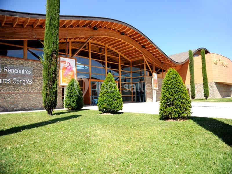 Façade d'un bâtiment moderne avec toit ondulé, grandes baies vitrées, pelouse et arbres en premier plan.
