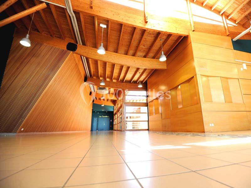 Intérieur d'un bâtiment avec structure en bois, plafond à poutres apparentes et sol carrelé éclairé par la lumière naturelle.