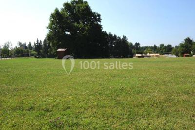 Vue d'une prairie verdoyante avec un grand arbre à gauche et des bâtiments ruraux sous un ciel dégagé.