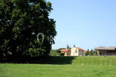 Vue d'une prairie verdoyante avec un grand arbre à gauche et des bâtiments ruraux sous un ciel dégagé.