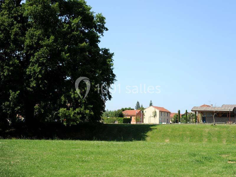 Vue d'une prairie verdoyante avec un grand arbre à gauche et des bâtiments ruraux sous un ciel dégagé.