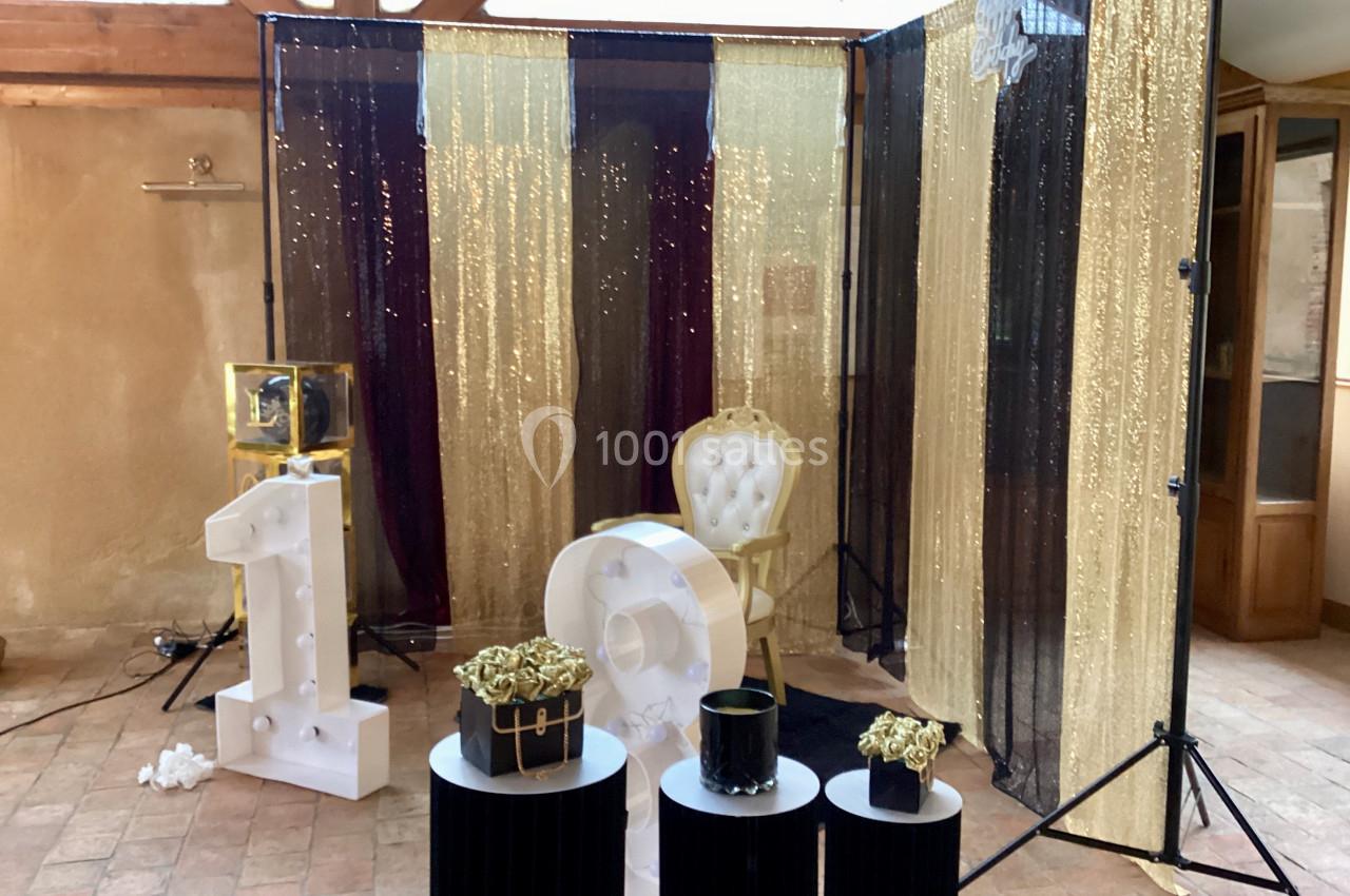 Décor festif avec rideaux dorés et noirs, fauteuil doré, chiffres ’18’ et petits présentoirs ornés de fleurs.