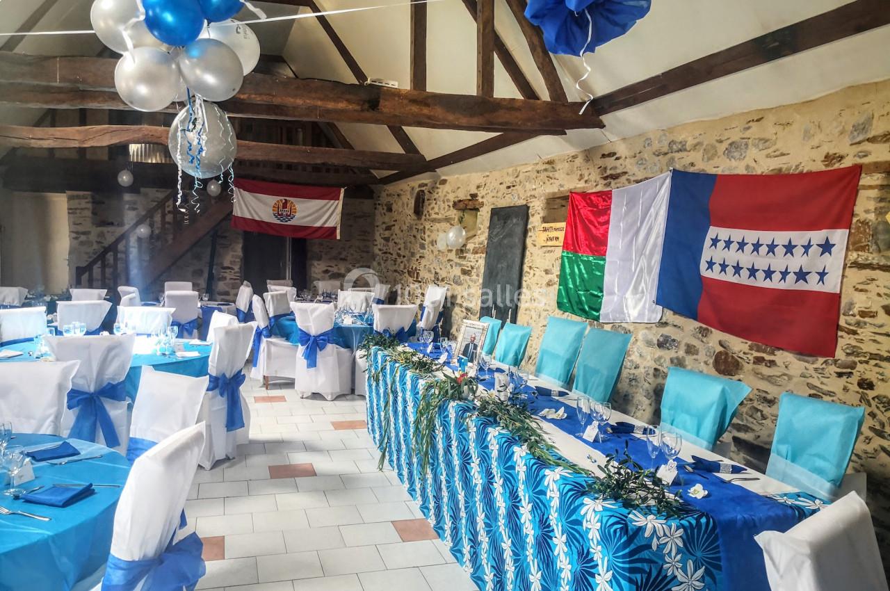 Salle décorée pour un événement avec des tables dressées, des nappes bleues, des drapeaux et des ballons suspendus.