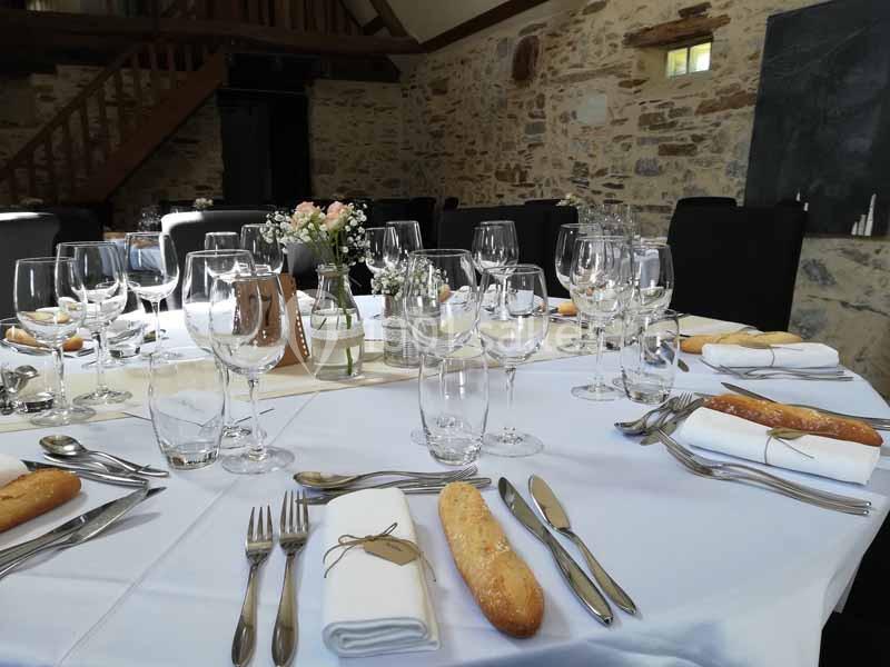 Table dressée avec nappes blanches, couverts, verres, fleurs et pains dans une salle en pierre avec poutres apparentes.