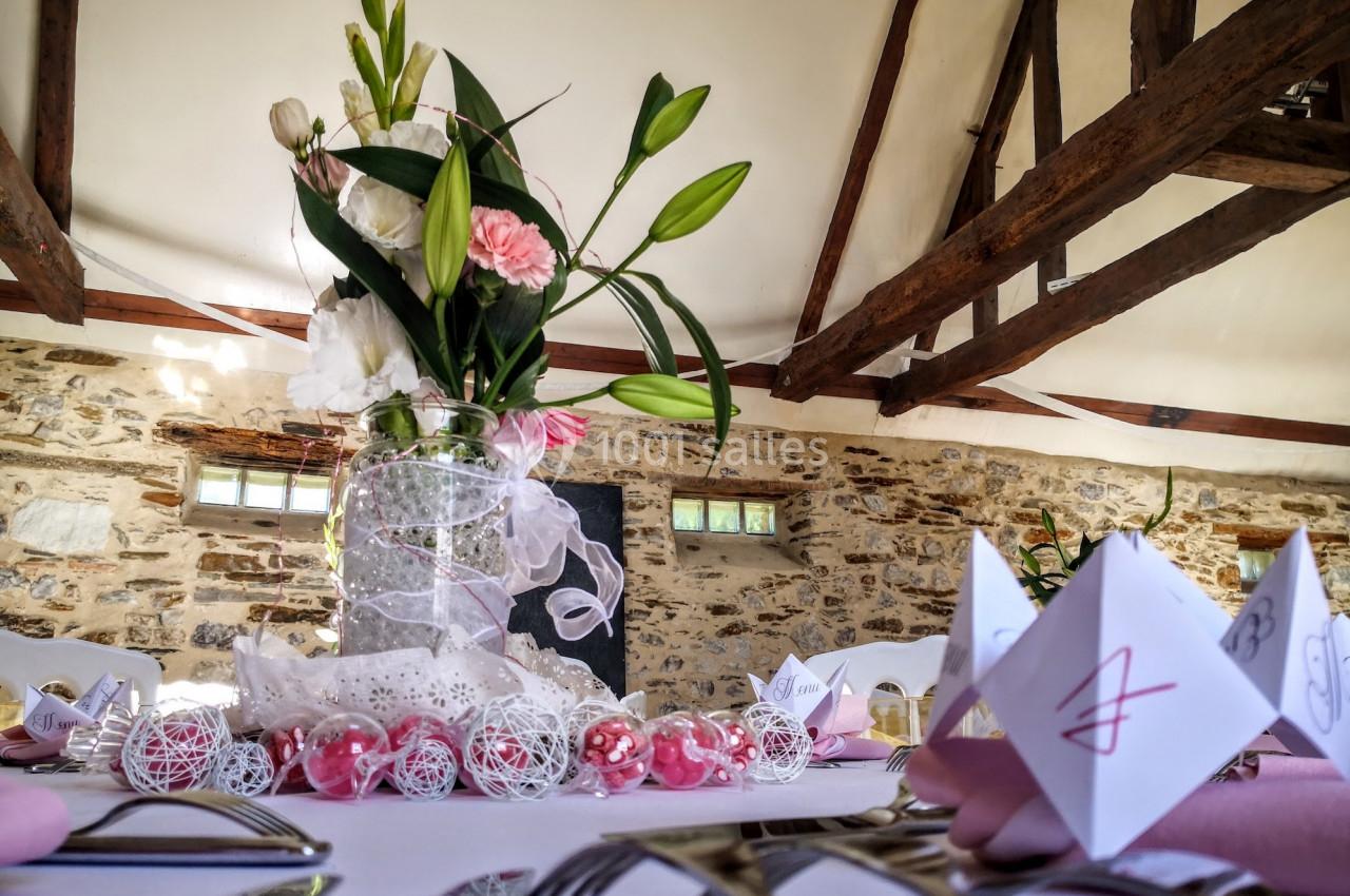 Centre de table floral avec des lys et des roses, décoré de rubans et perles, dans une salle aux poutres apparentes.