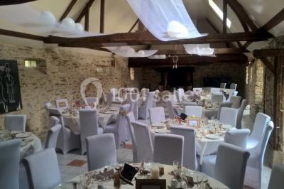 Salle à manger rustique avec table en bois dressée, mur en pierre apparente, étagères intégrées et cuisine ouverte.