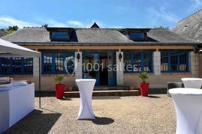 Salle à manger rustique avec table en bois dressée, mur en pierre apparente, étagères intégrées et cuisine ouverte.