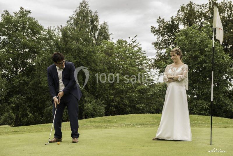 Un homme en costume joue au golf sur un green, observé par une femme en robe de mariée, bras croisés.