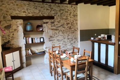 Salle à manger rustique avec table en bois dressée, mur en pierre apparente, étagères intégrées et cuisine ouverte.