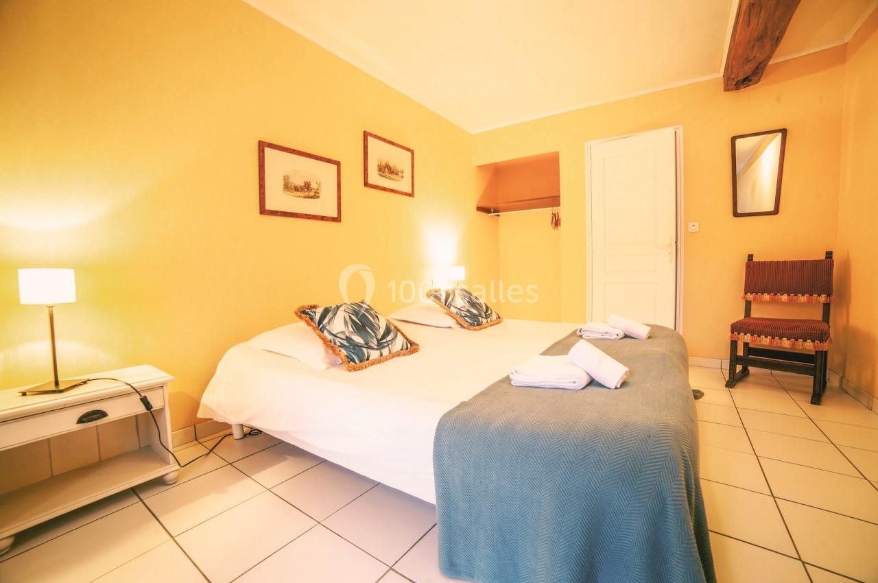 Chambre lumineuse avec lit double, draps bleus, lampes de chevet, murs jaunes et poutre apparente au plafond.