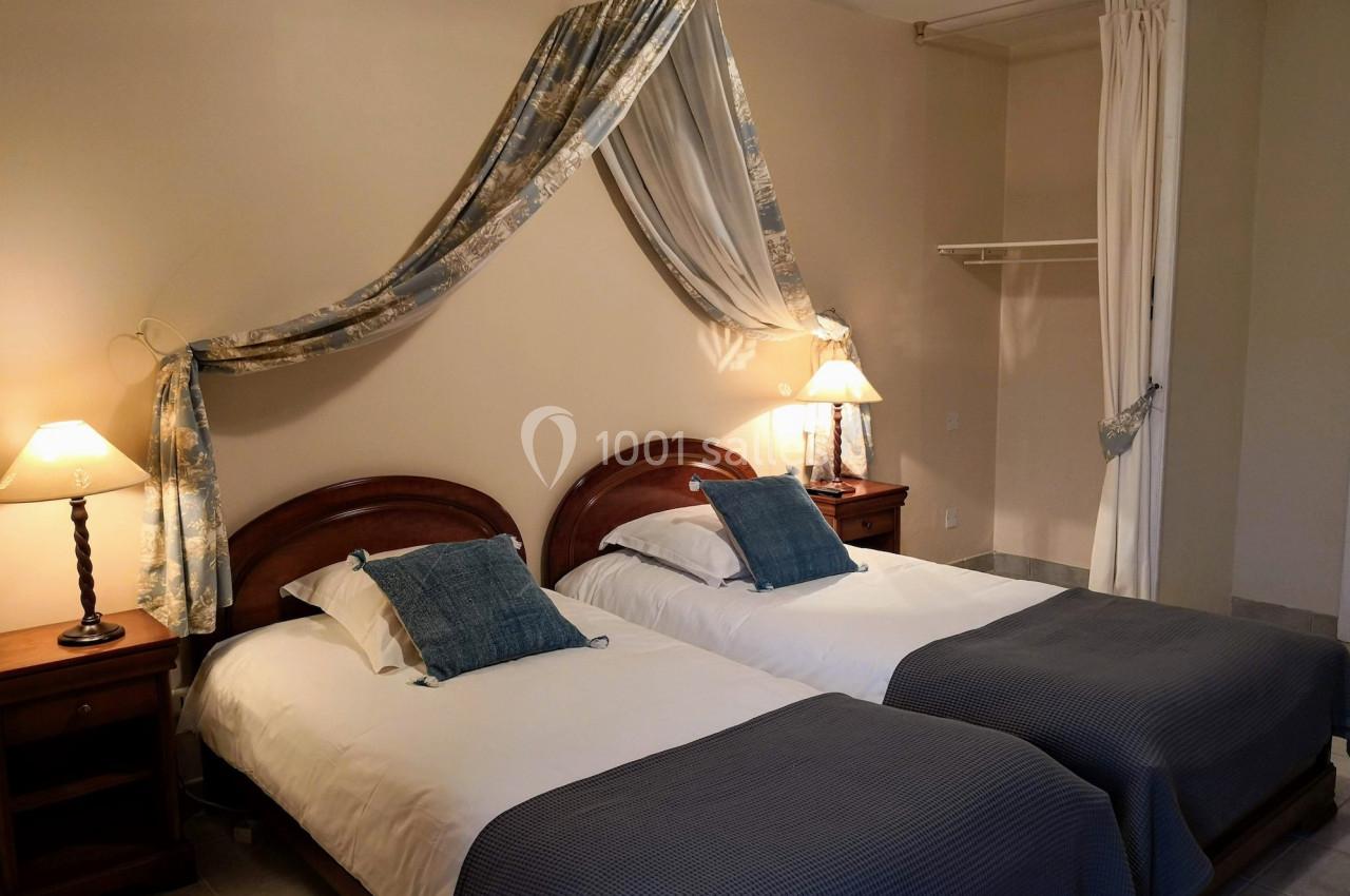 Deux lits simples avec couvre-lits bleus, encadrés par des tables de chevet et éclairés par des lampes, dans une chambre…