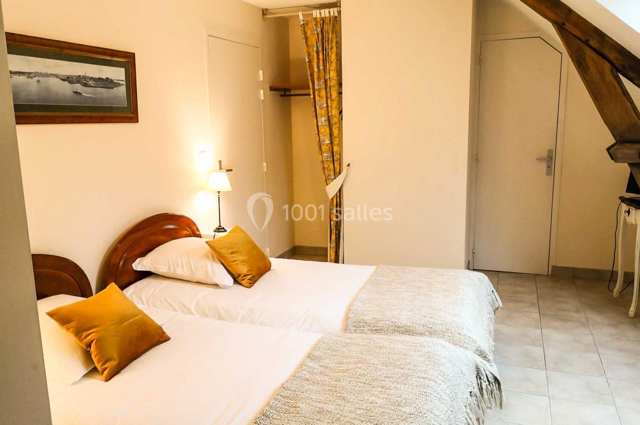 Deux lits simples avec coussins jaunes dans une chambre lumineuse, décorée avec une poutre apparente et un rideau.