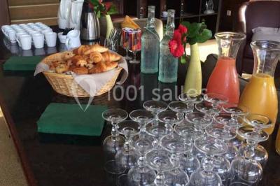 Salle à manger rustique avec table en bois dressée, mur en pierre apparente, étagères intégrées et cuisine ouverte.