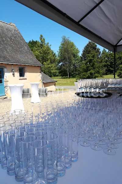 Verres en verre alignés sur une table sous une tente, avec un bâtiment en pierre et un jardin en arrière-plan.
