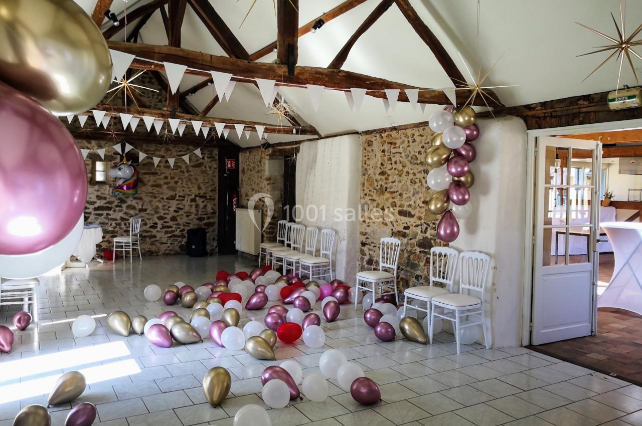 Salle décorée pour une fête avec des ballons colorés au sol, des guirlandes et des chaises alignées contre un mur en pierre.