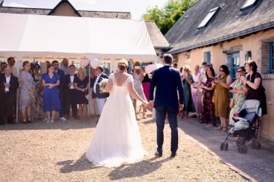 Salle à manger rustique avec table en bois dressée, mur en pierre apparente, étagères intégrées et cuisine ouverte.