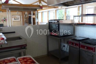 Salle à manger rustique avec table en bois dressée, mur en pierre apparente, étagères intégrées et cuisine ouverte.