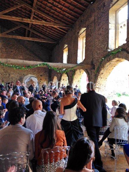 Une mariée et son accompagnateur avancent dans une salle en pierre remplie d'invités assis sur des chaises transparentes.