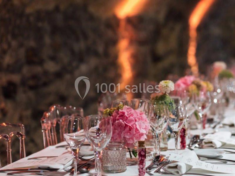 Table élégamment dressée avec des fleurs roses, verres et couverts, dans un cadre aux murs en pierre éclairés.