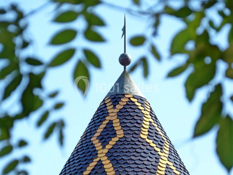 Toit conique recouvert d'ardoises colorées avec un motif géométrique, surmonté d'une flèche métallique, encadré de feuillage.