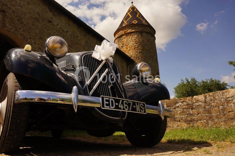 Voiture ancienne noire décorée d'un nœud blanc, garée devant une tour en pierre sous un ciel partiellement nuageux.