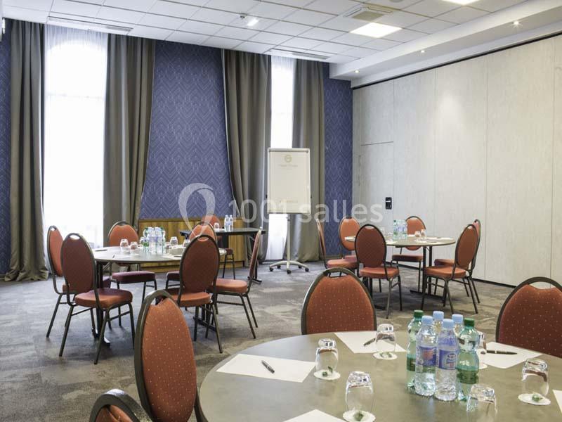 Salle de réunion lumineuse avec tables rondes, chaises rouges, tableau blanc et bouteilles d'eau disposées.