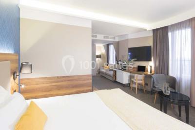 Chambre d'hôtel lumineuse avec lit double, coin salon, télévision murale et grandes fenêtres avec rideaux.