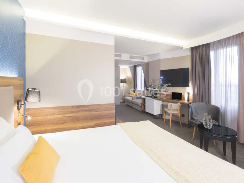 Chambre d'hôtel lumineuse avec lit double, coin salon, télévision murale et grandes fenêtres avec rideaux.