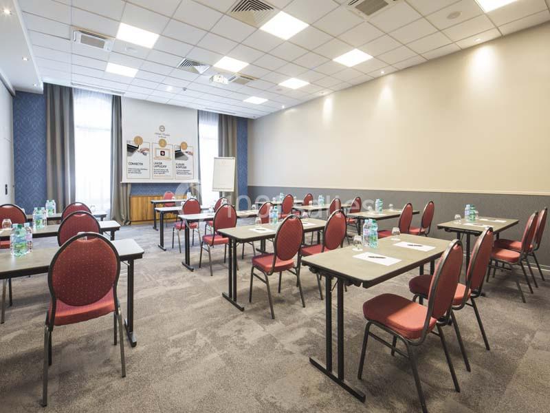 Salle de réunion lumineuse avec des tables alignées, des chaises rouges et des bouteilles d'eau sur chaque table.