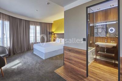 Chambre d'hôtel lumineuse avec lit double, coin salon, télévision murale et grandes fenêtres avec rideaux.