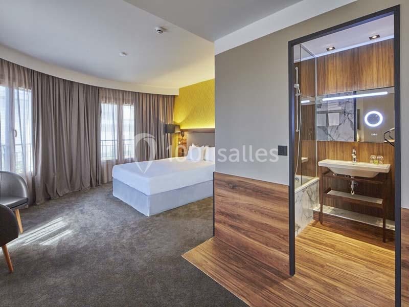 Chambre d'hôtel moderne avec lit double, grandes fenêtres, rideaux opaques et salle de bain ouverte avec éclairage…