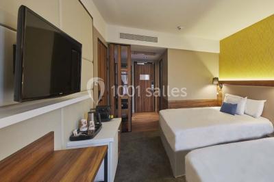 Chambre d'hôtel lumineuse avec lit double, coin salon, télévision murale et grandes fenêtres avec rideaux.