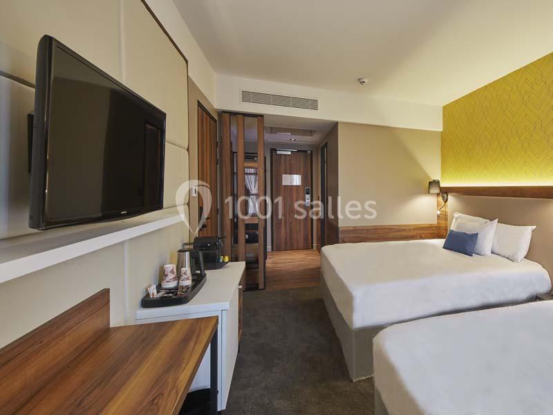 Chambre d'hôtel moderne avec deux lits, télévision murale, bureau en bois et éclairage chaleureux.