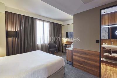 Chambre d'hôtel lumineuse avec lit double, coin salon, télévision murale et grandes fenêtres avec rideaux.