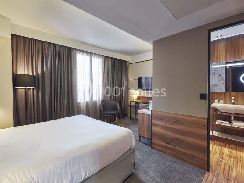 Chambre d'hôtel moderne avec lit double, bureau, télévision, éclairage tamisé et salle de bain attenante visible.