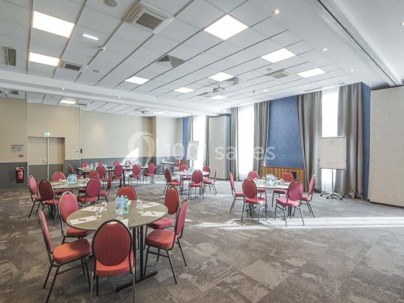 Salle de réunion lumineuse avec plusieurs tables rondes, chaises rouges et équipements de présentation.