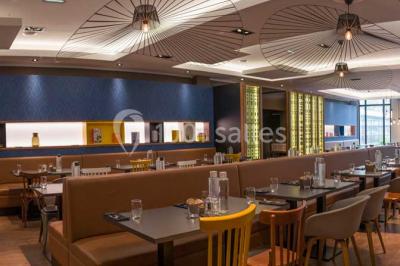 Chambre d'hôtel lumineuse avec lit double, coin salon, télévision murale et grandes fenêtres avec rideaux.