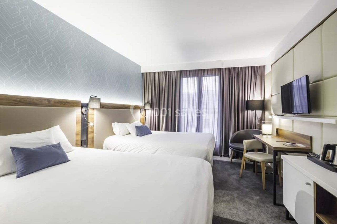 Chambre d'hôtel moderne avec deux lits, bureau, télévision murale et grandes fenêtres avec rideaux.