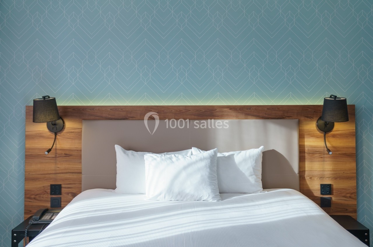 Lit double avec tête de lit en bois, deux lampes murales noires et un mur bleu à motifs géométriques en arrière-plan.