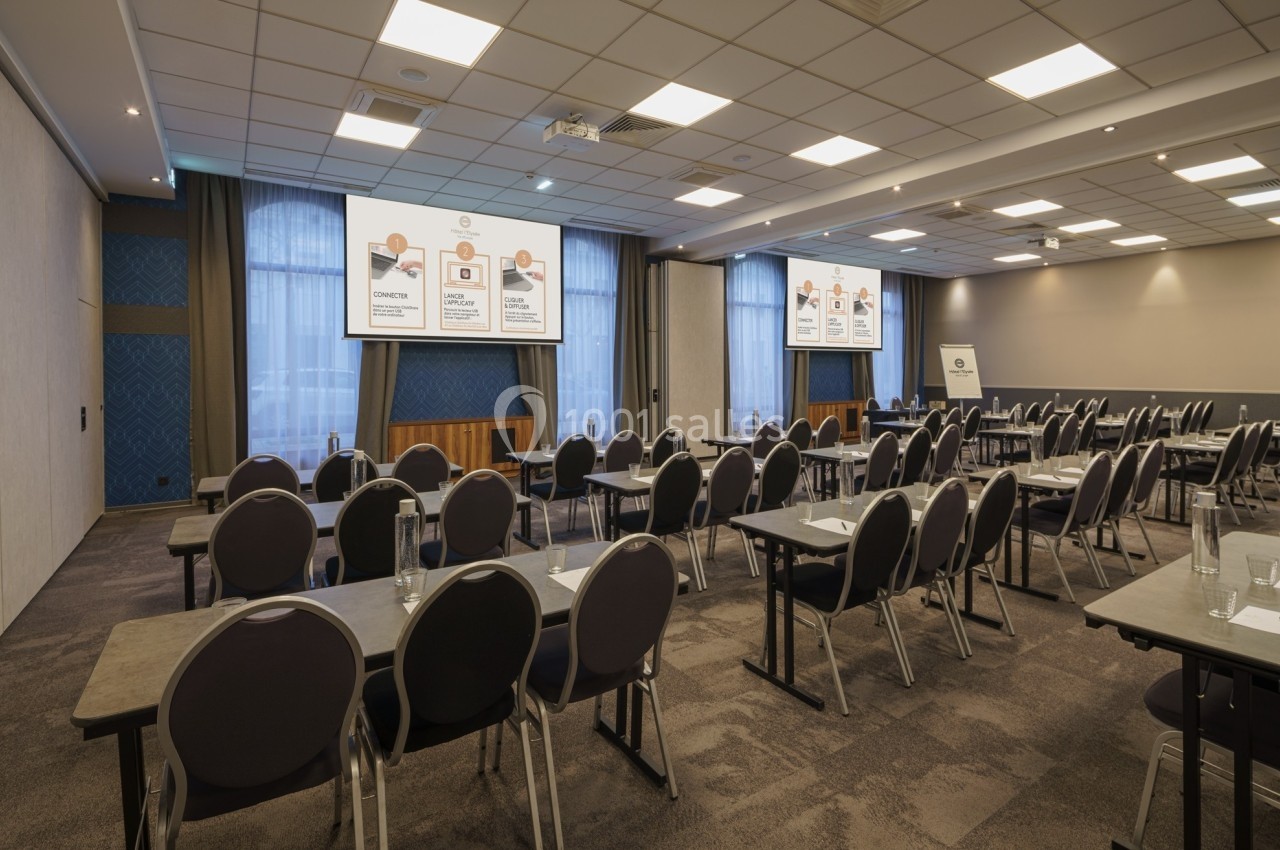 Salle de conférence vide avec des rangées de chaises, des tables et des écrans affichant des présentations.