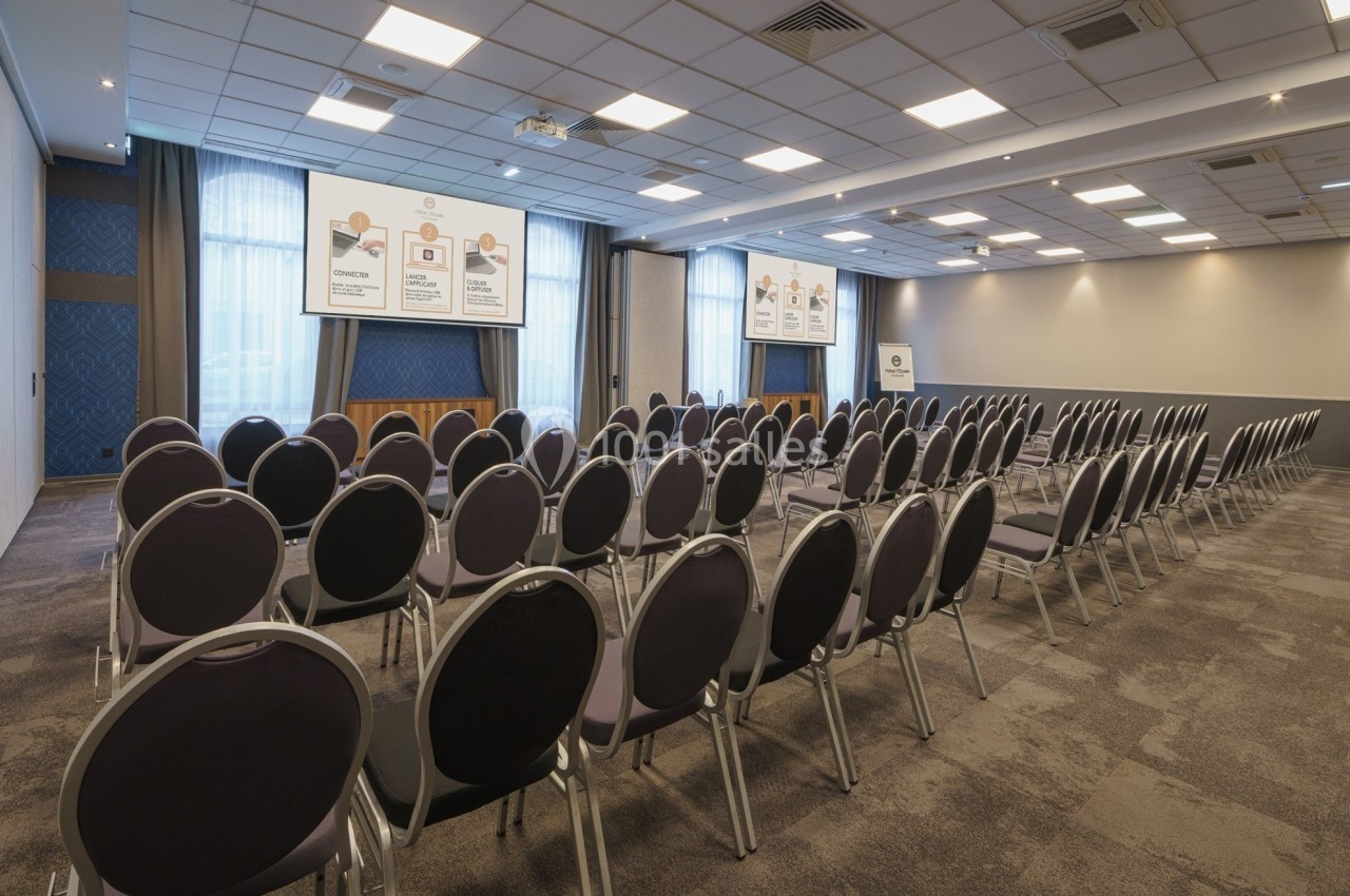 Salle de conférence vide avec des rangées de chaises alignées face à trois écrans de projection allumés.