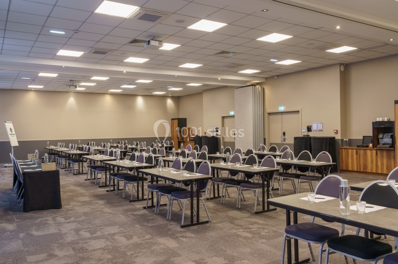 Salle de conférence vide avec des tables et des chaises alignées, éclairée par des plafonniers.