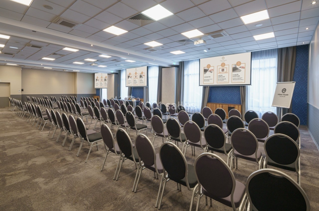Salle de conférence vide avec des rangées de chaises noires, un écran de projection et des affiches explicatives au mur.