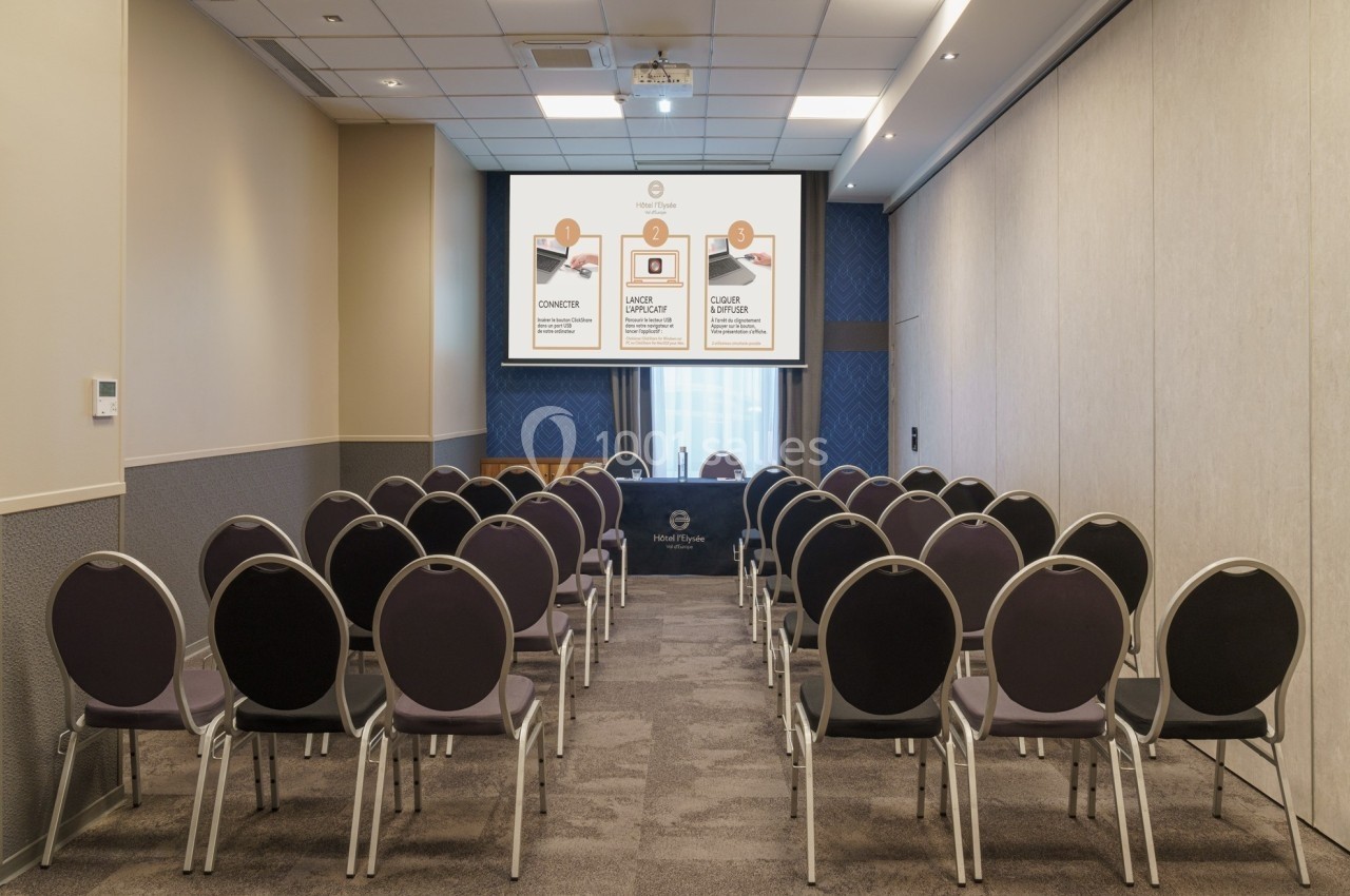 Salle de conférence vide avec des rangées de chaises alignées face à un écran de projection allumé.