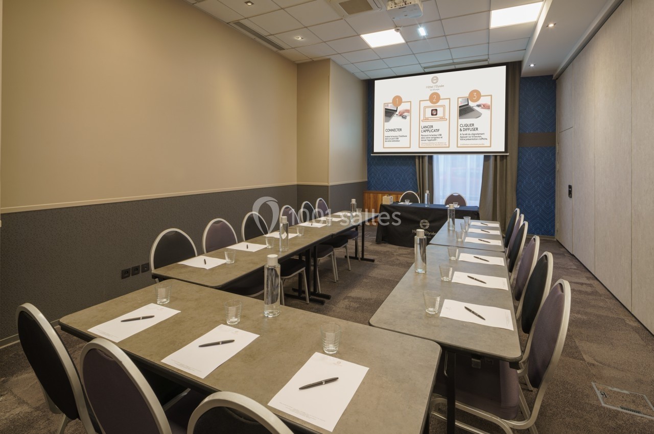 Salle de réunion équipée de tables en U, chaises, blocs-notes, bouteilles d'eau et écran de présentation allumé.