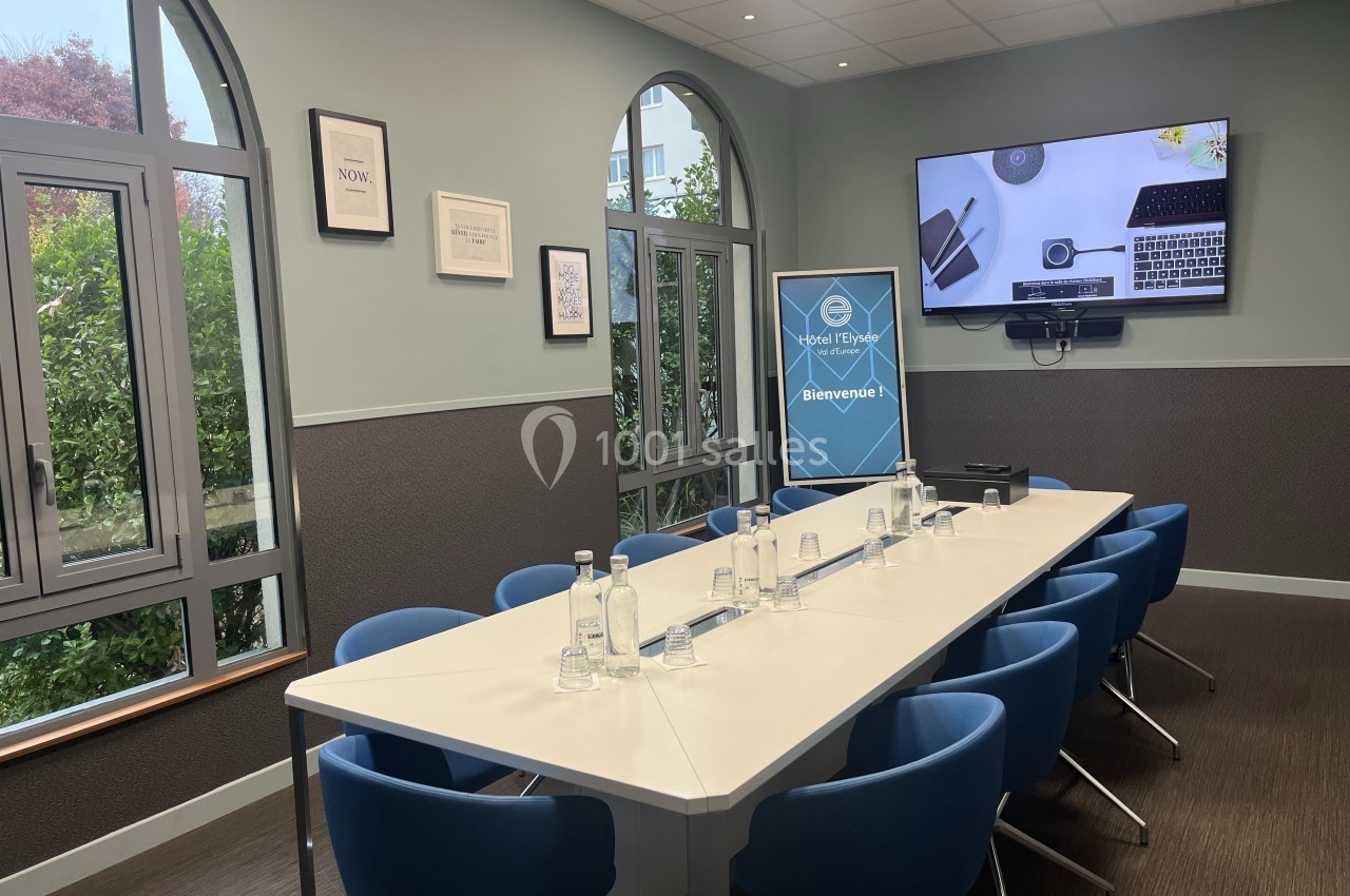 Salle de réunion lumineuse avec table blanche, chaises bleues, écran mural et fenêtres donnant sur un jardin.