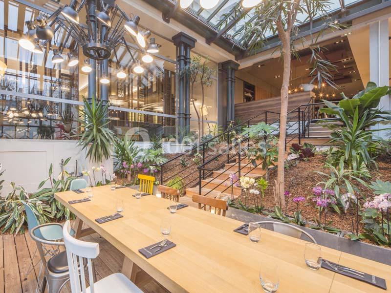 Salle lumineuse avec grande table en bois, chaises dépareillées, plantes tropicales et verrière laissant entrer la lumière…