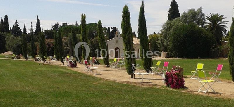 Allée bordée de cyprès avec des chaises colorées et des jardinières fleuries, menant à une petite chapelle.