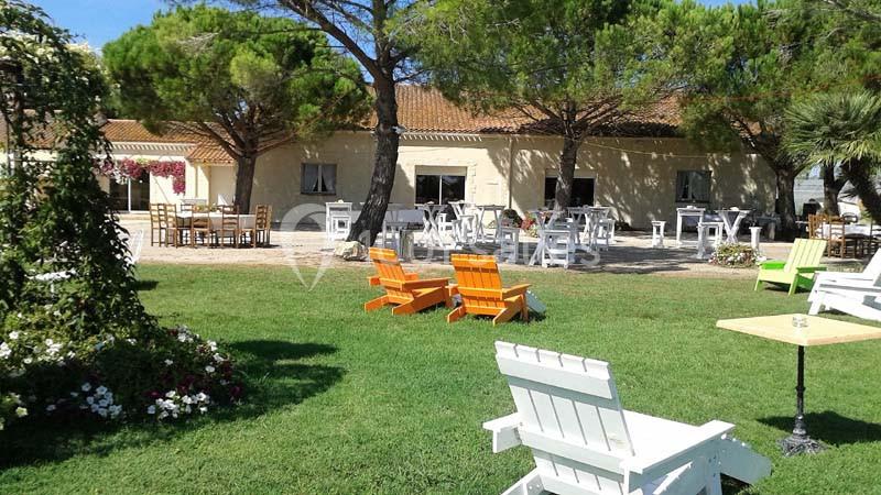 Espace extérieur avec pelouse, chaises colorées, tables et arbres, devant un bâtiment en pierre sous un ciel dégagé.