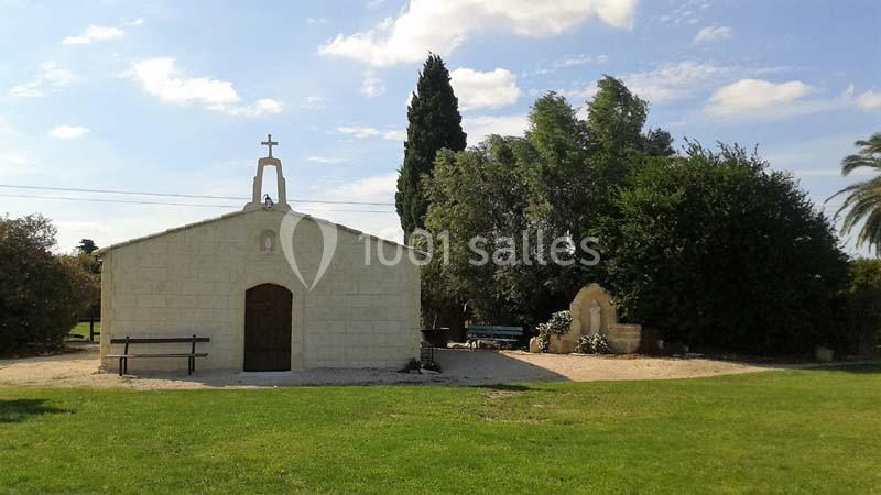 Petite chapelle en pierre entourée d'arbres et d'un espace vert sous un ciel partiellement nuageux.